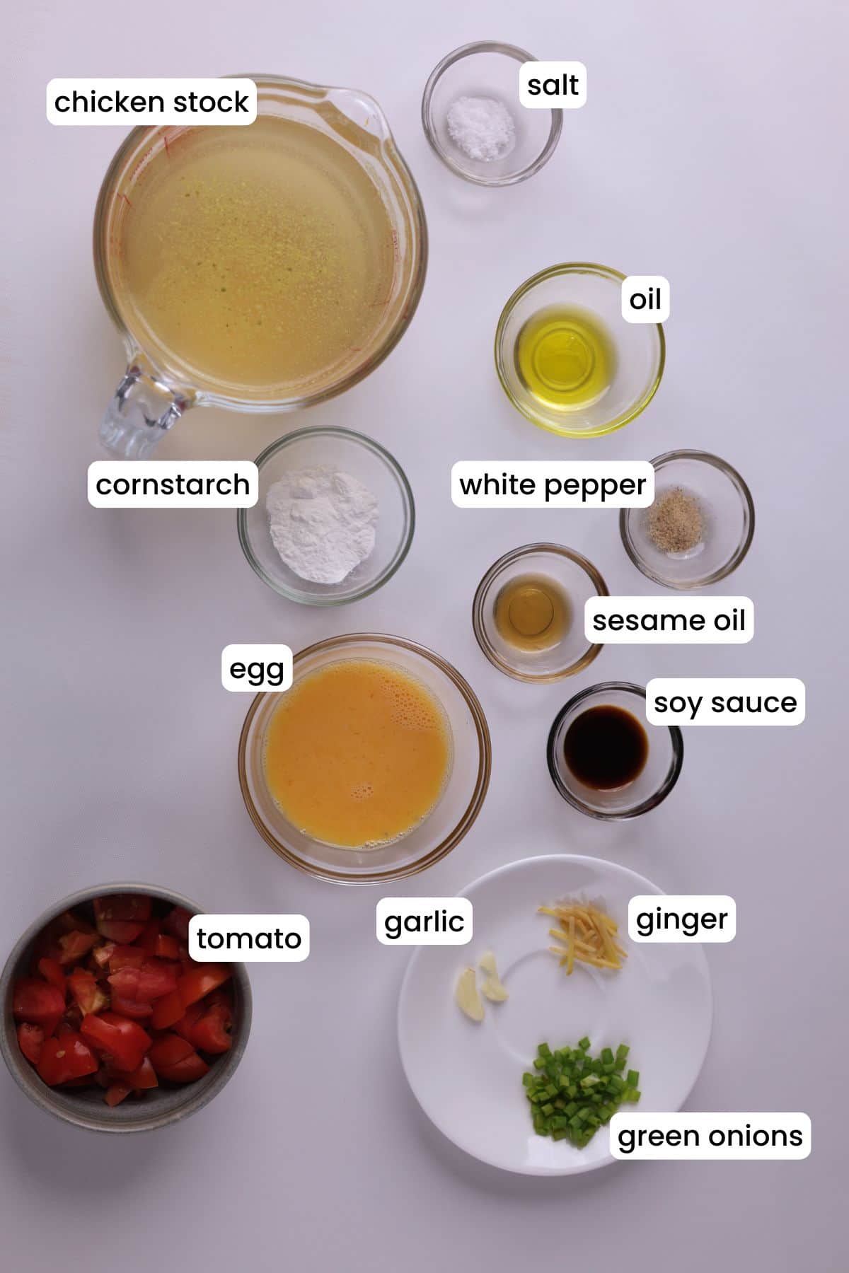 All ingredients for tomato egg drop soup arranged on a white surface, including tomatoes, eggs, green onions, ginger, garlic, chicken stock, soy sauce, sesame oil, and cornstarch.