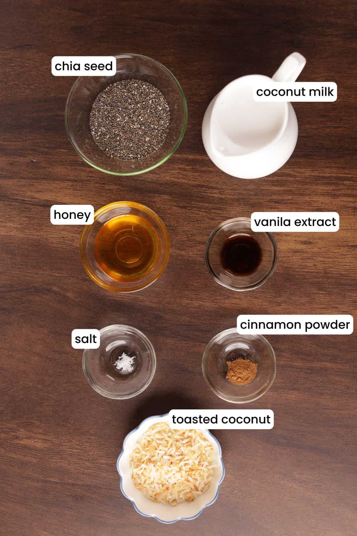 Ingredients for coconut chia seed pudding arranged on a wooden background, including chia seeds, coconut milk, honey, vanilla, cinnamon, salt, and toasted coconut.