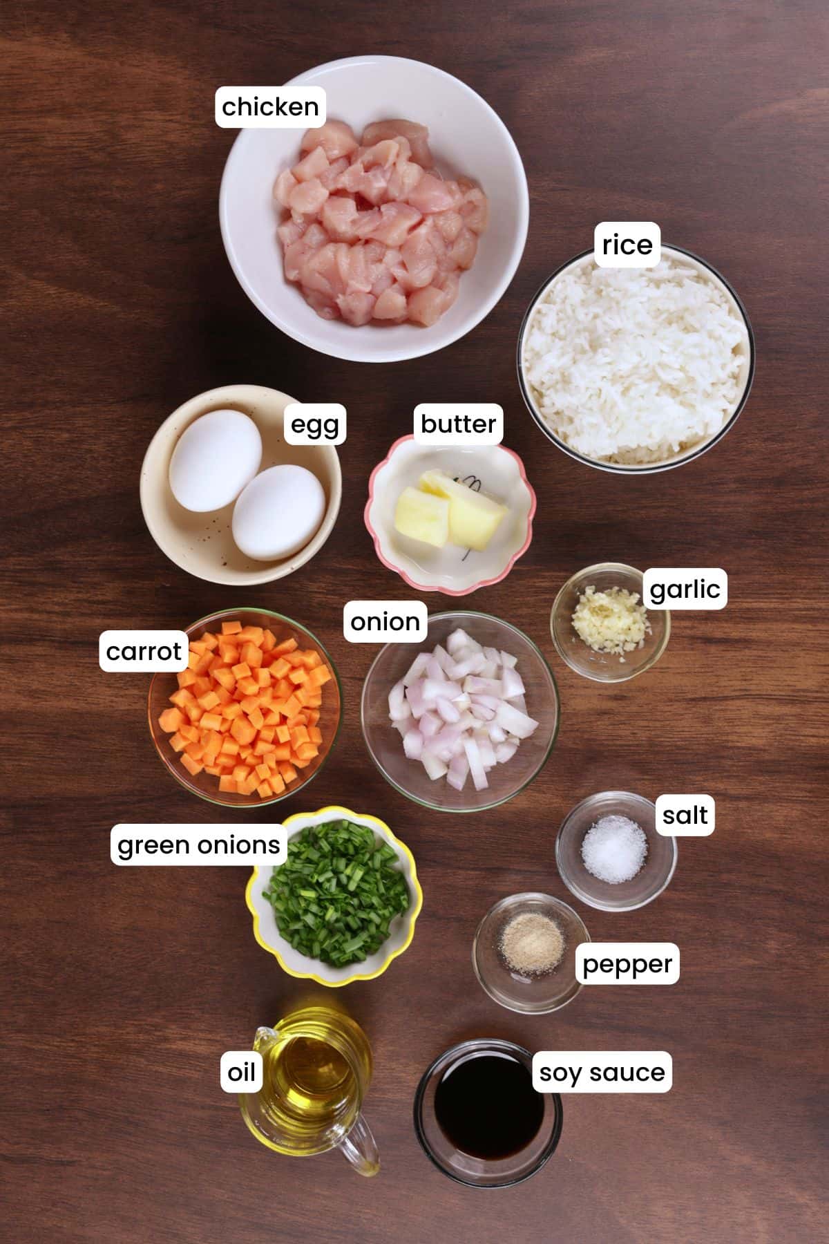 Ingredients for hibachi chicken fried rice laid out on a wooden background including rice, chicken, eggs, garlic, soy sauce, carrot, onion, and butter.