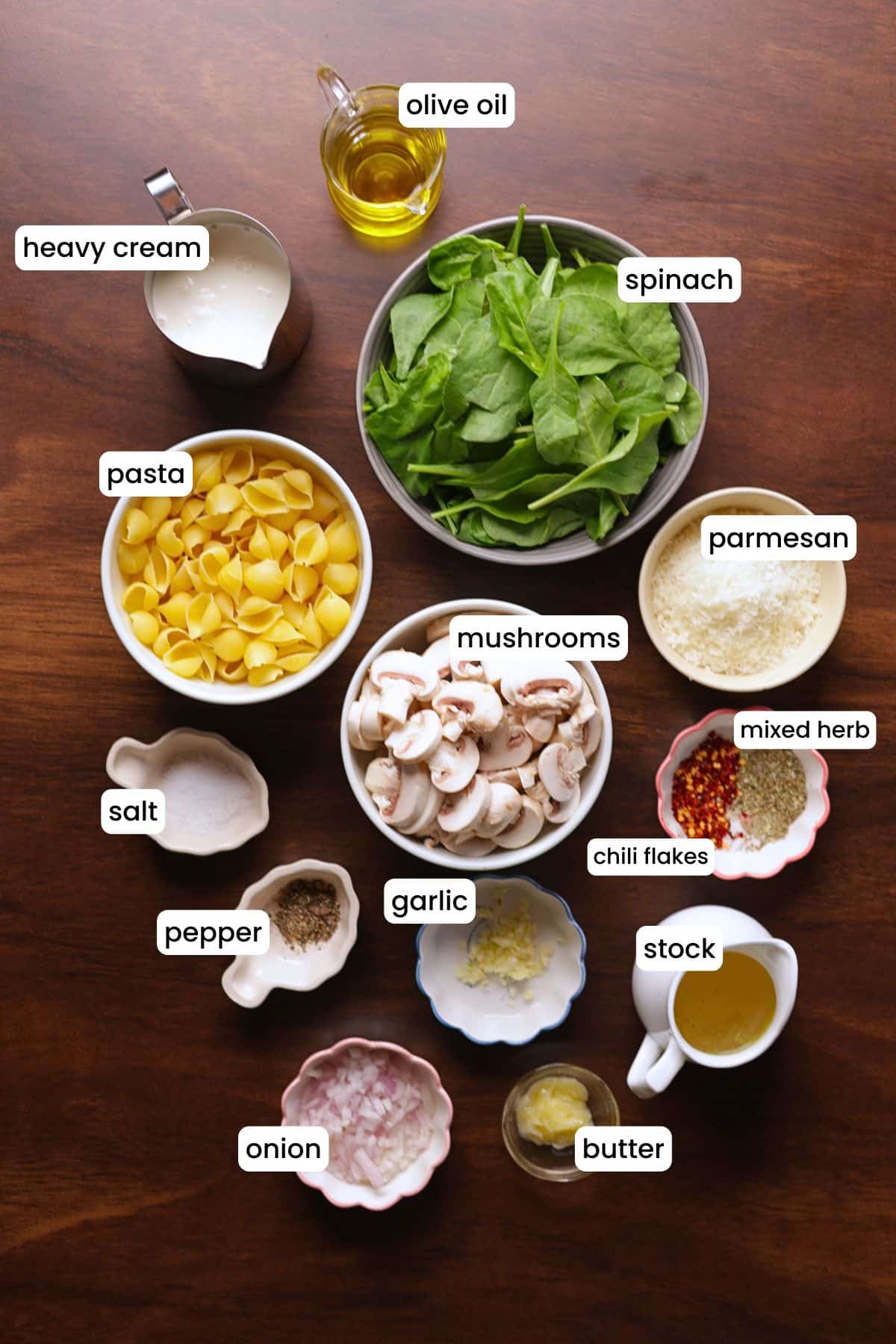 All ingredients of spinach mushroom pasta are arranged on a wooden background, including mushrooms, spinach, pasta shells, garlic, onion, olive oil, butter, chili flakes, Italian seasoning, stock, cream, and Parmesan cheese.