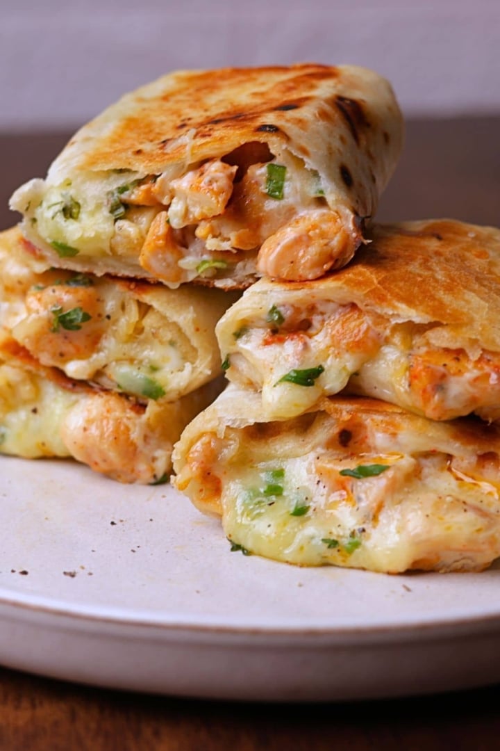 Melty cheesy chicken wraps stack on a off white platter.