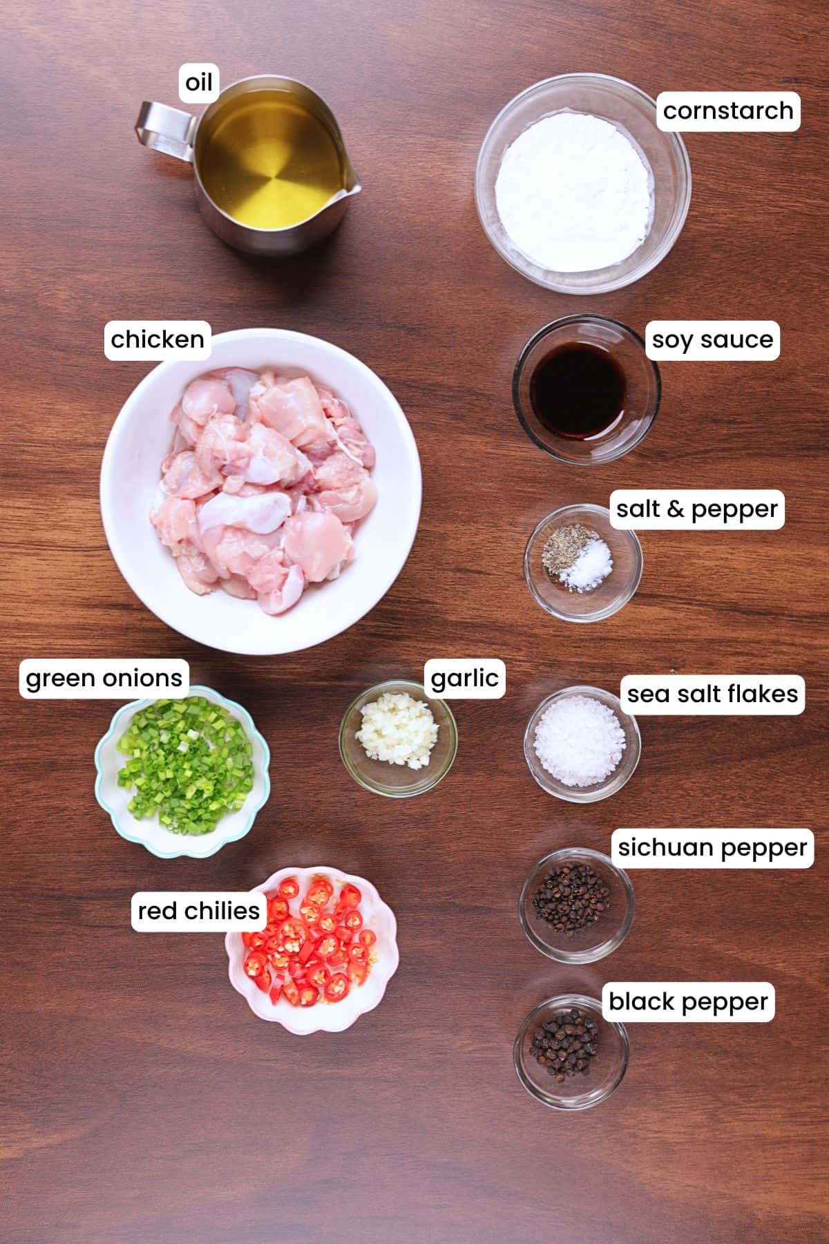Salt and pepper chicken ingredients such as chicken, garlic, red chilies, sichuan pepper, black pepper, salt, oil, and green onions are placed on a wooden backdrop.